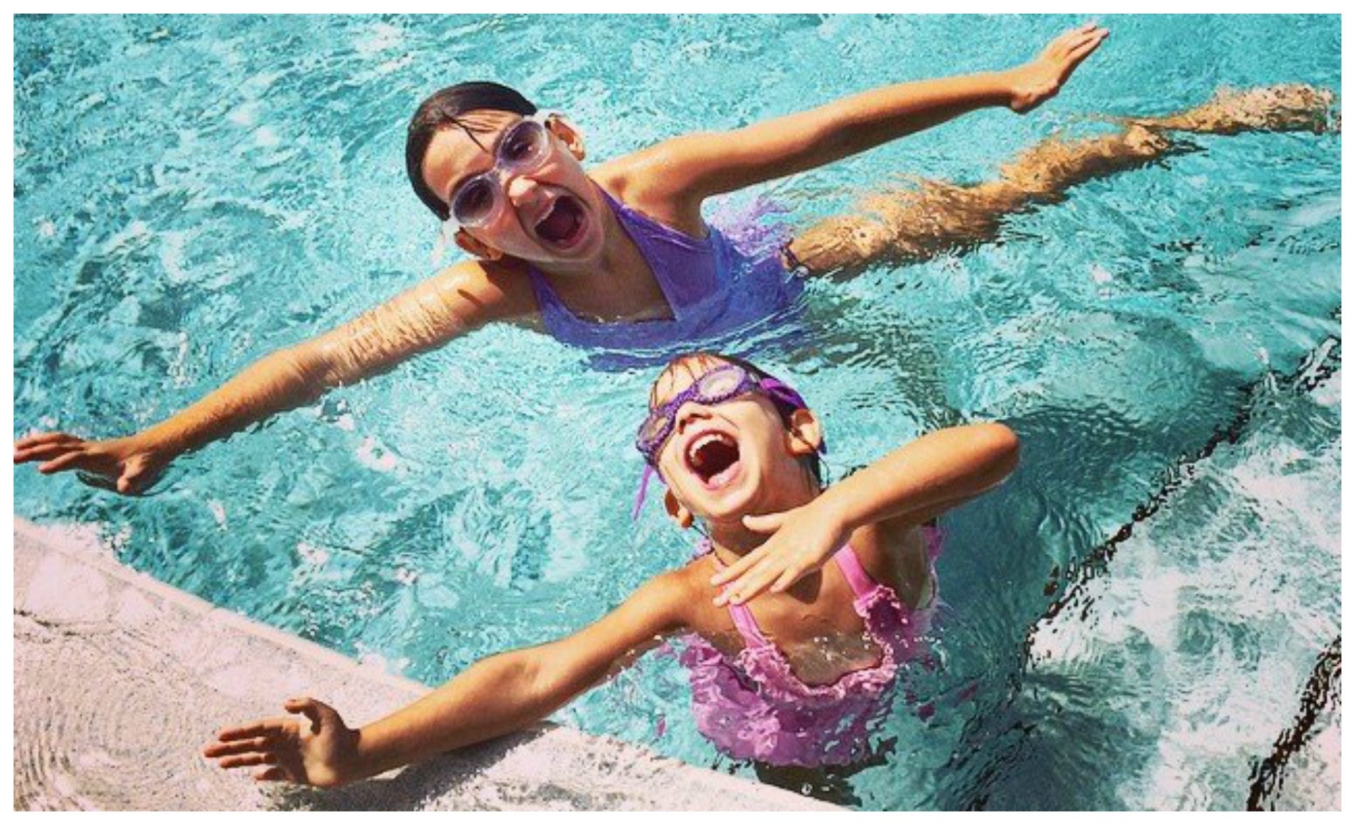 Two adults in swimwear and goggles playfully shout and flail their arms in a swimming pool.