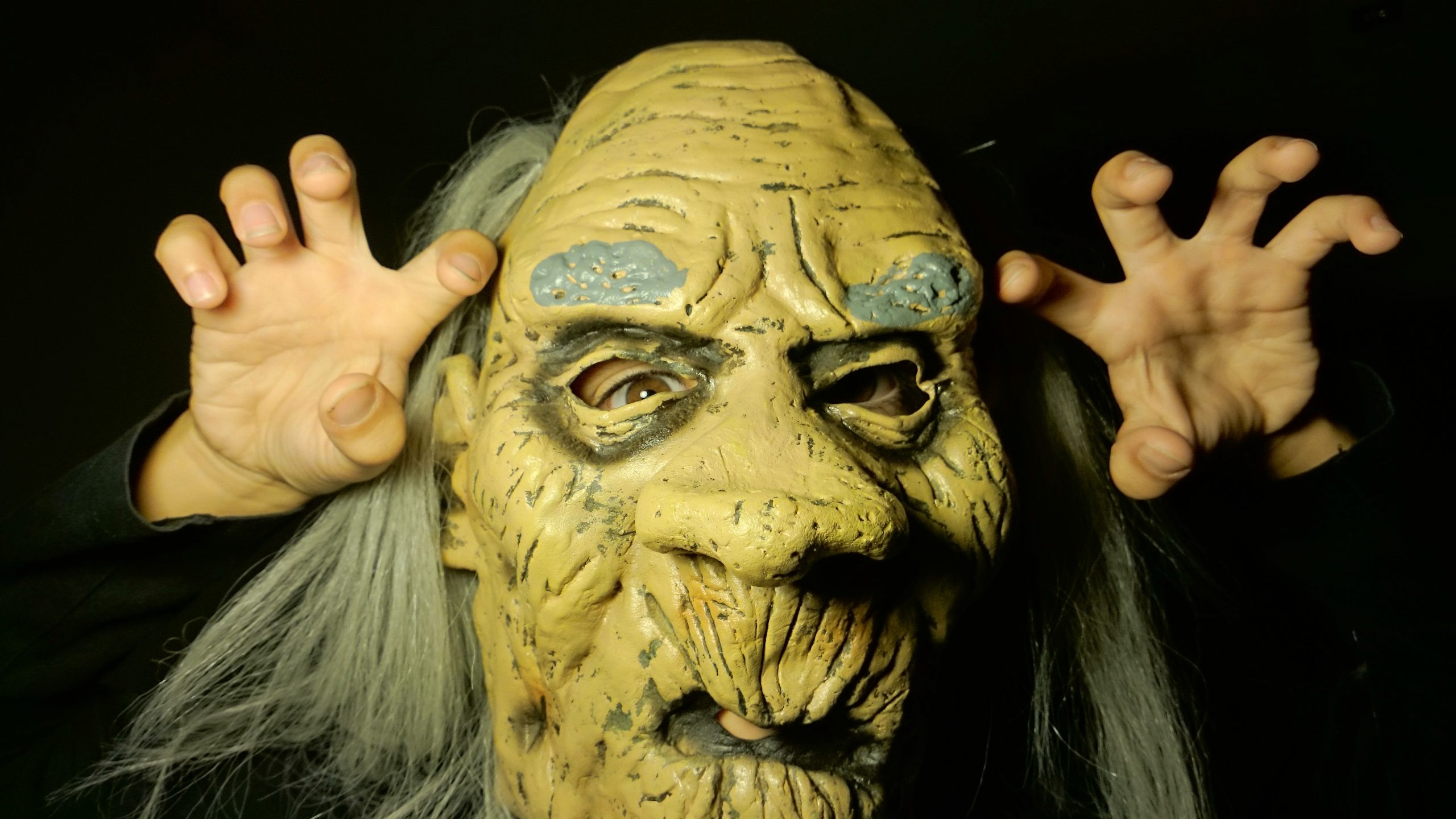Person wearing a scary monster mask with long grey hair, making claw-like gestures with their hands in anger.