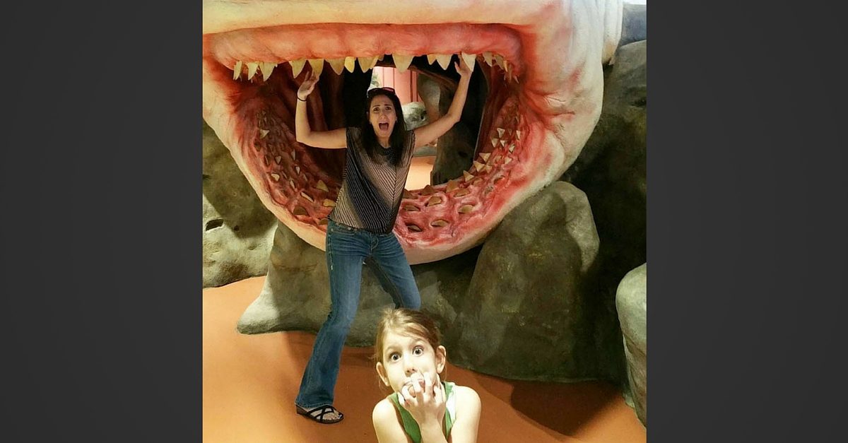 Woman playing playfully inside a large, model dinosaur mouth with a child crouching in front.